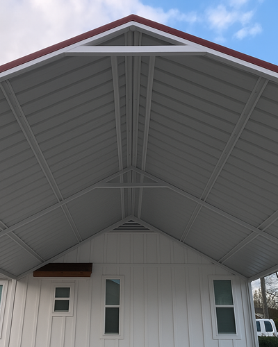 Upward view of a gable-style metal carport with red trim, white support beams, and a white exterior wall featuring small rectangular windows.
